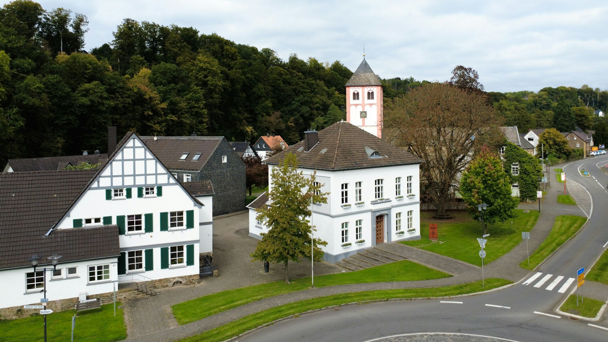 Luftbild von zwei weißen Häusern an einem Kreisverkehr, dahinter ein Kirchturm.