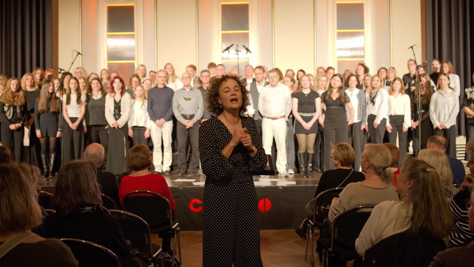 Die Chorleiterin steht im Gang zwischen den Sitzreihen, im Hintergrund der Chor auf der Bühne.
