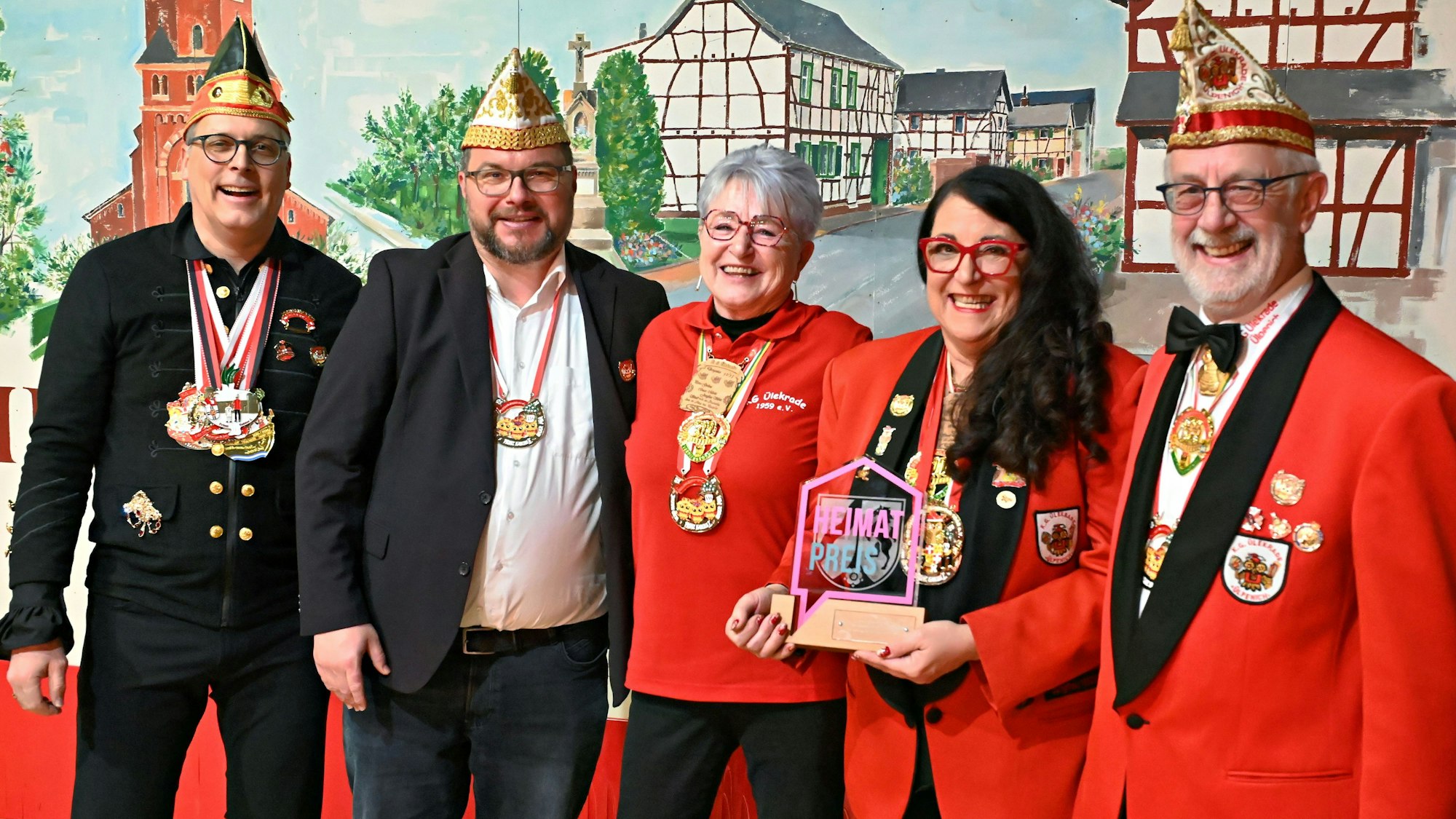 Drei Männer und zwei Frauen stehen mit dem Preis vor einem Bühnenbild, das den Ortskern von Ülpenich zeigt.