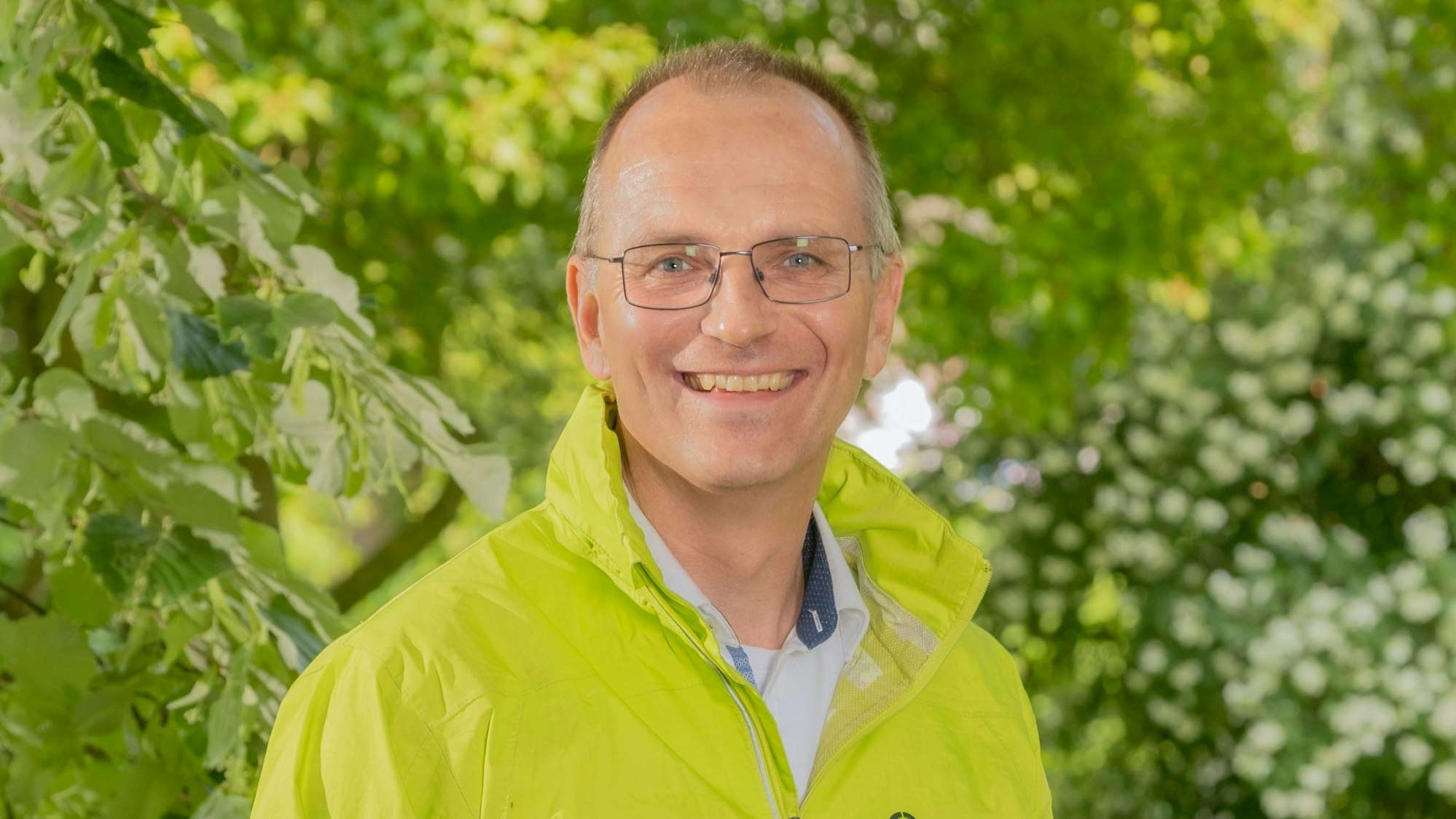 Dirk Jansen trägt eine Brille und eine hellgrüne Jacke. Er lächelt in die Kamera. Im Hintergrund Bäume und Büsche.