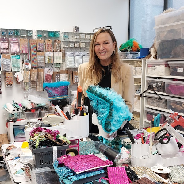 Eine Frau in einem Schneideratelier. Sie hat einen bunten Karnevalshut mit Federn in der Hand.