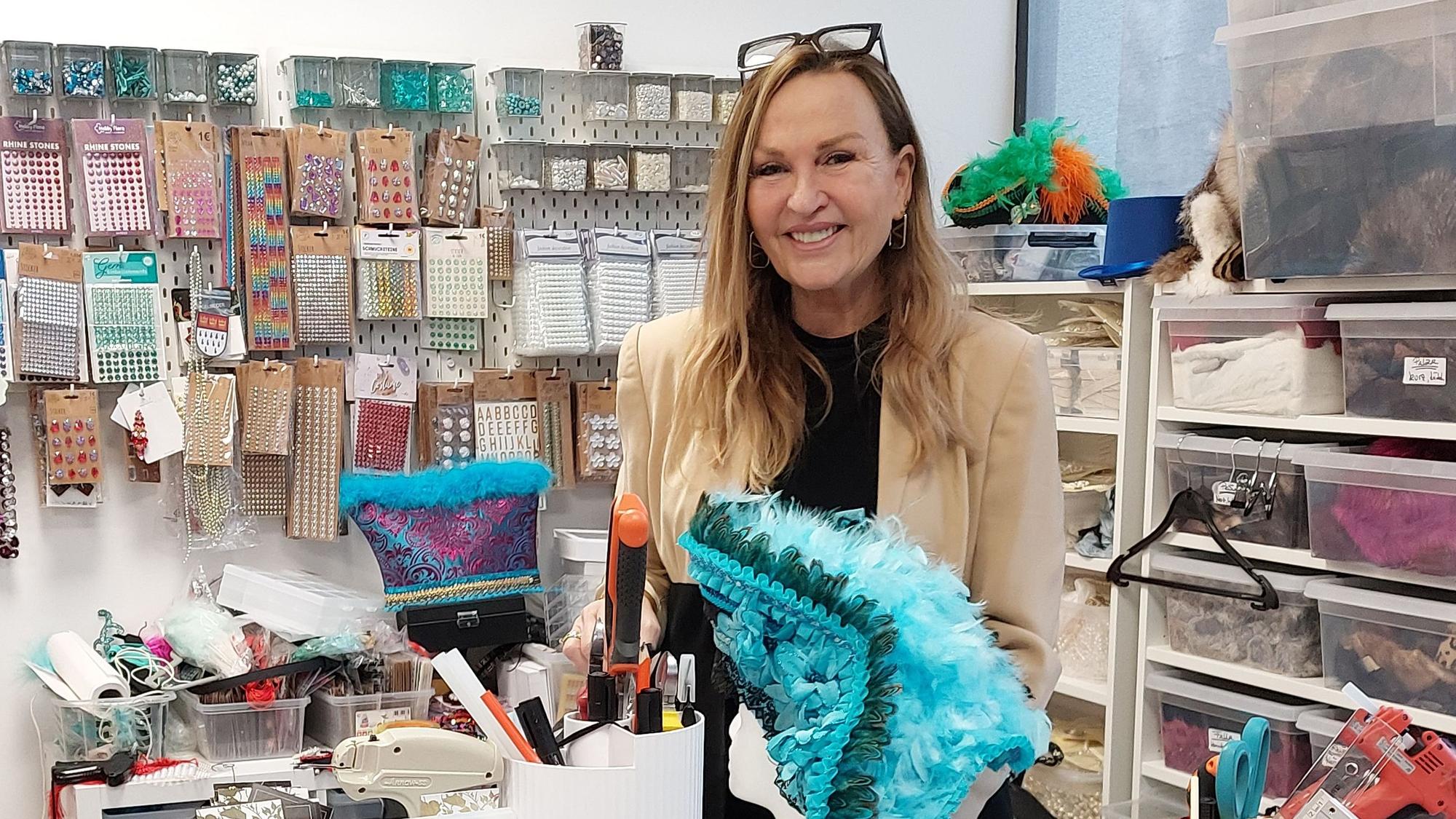 Eine Frau in einem Schneideratelier. Sie hat einen bunten Karnevalshut mit Federn in der Hand.