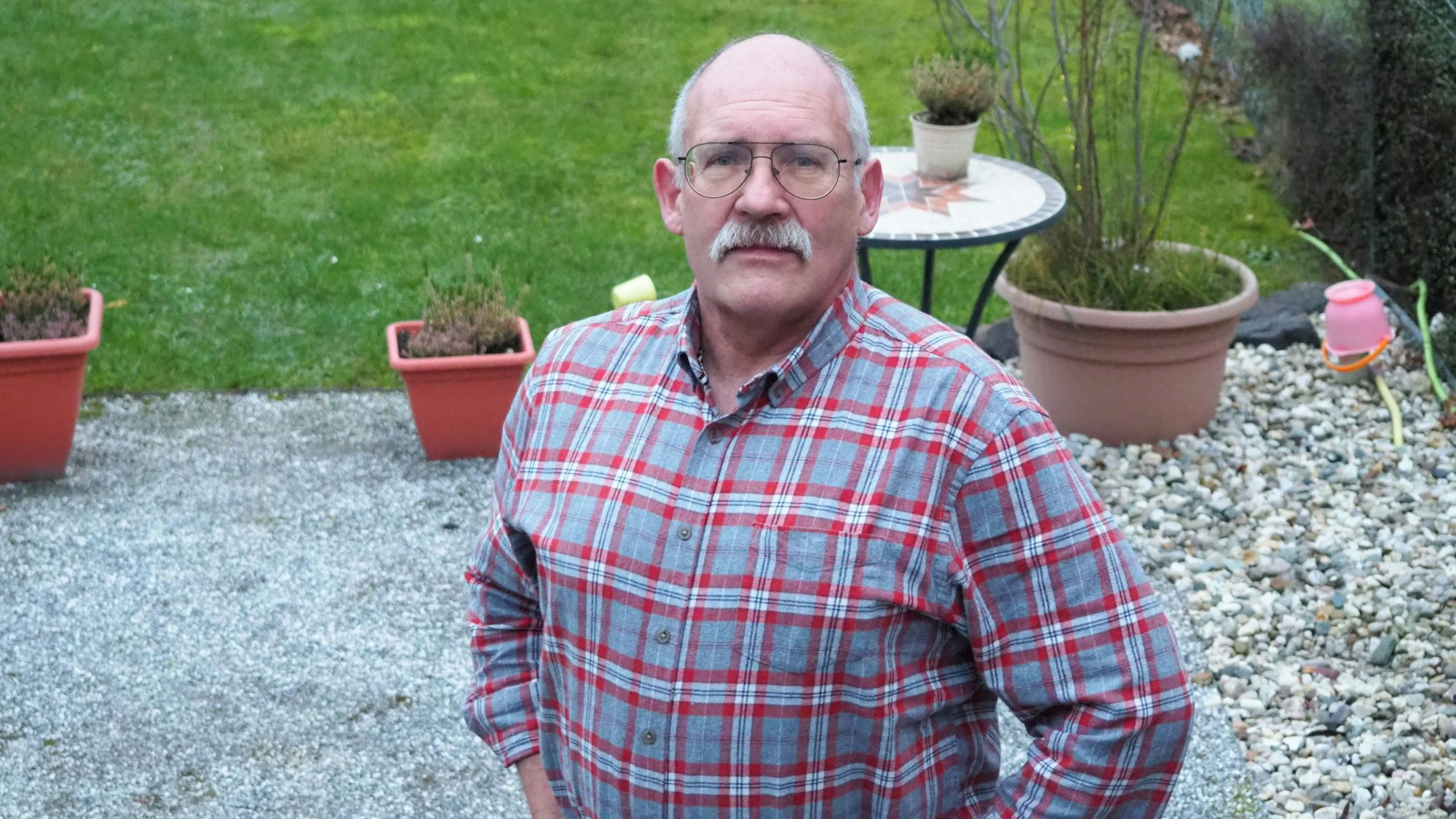 Thomas Enke, Stirnglatze, Schnurrbart, kariertes Hemd, steht in einem Garten.