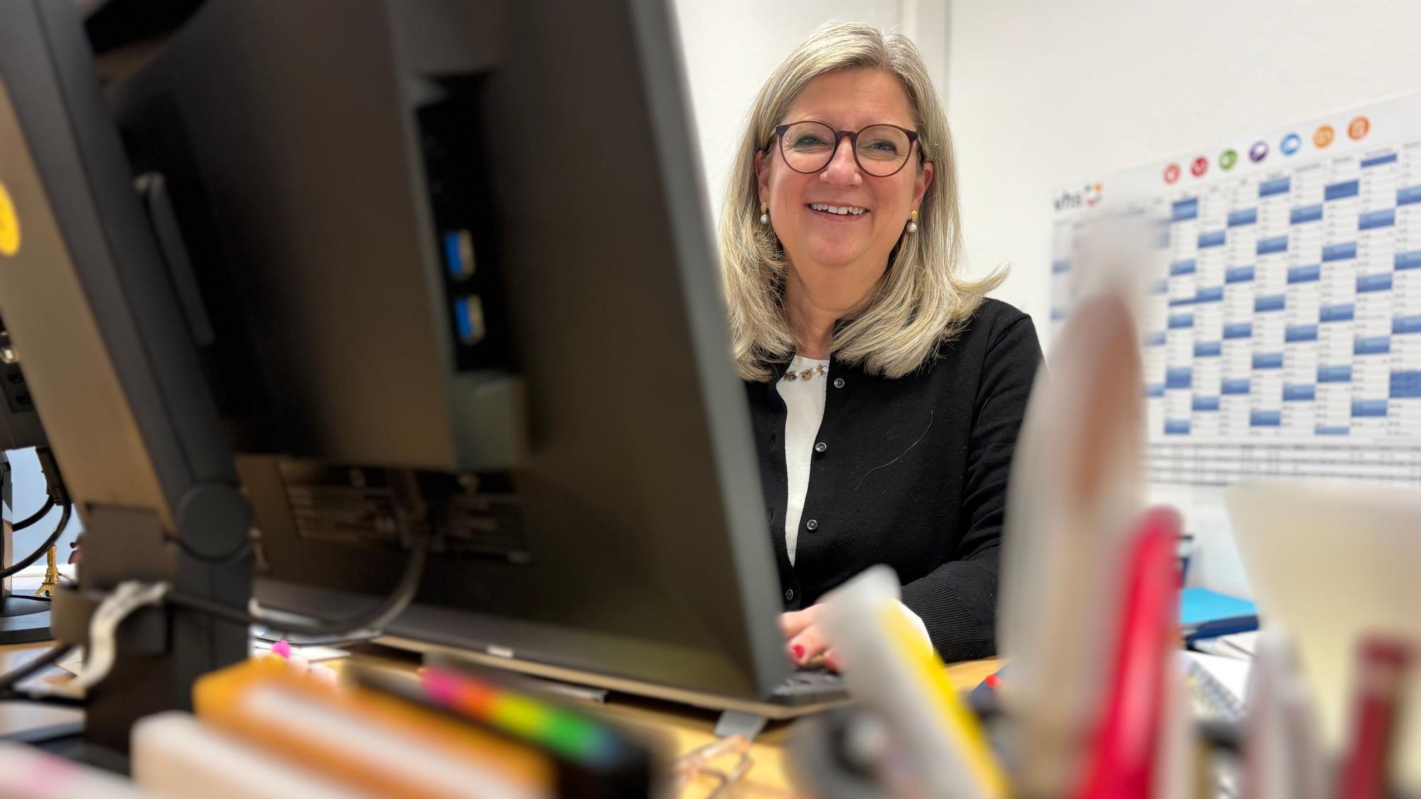 Vanessa Becker sitzt hinter einem Computerbildschirm an einem Schreibtisch und lächelt. Im Vordergrund sind unscharfe Büroartikel.