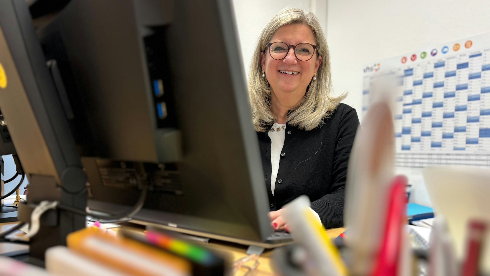 Vanessa Becker sitzt hinter einem Computerbildschirm an einem Schreibtisch und lächelt. Im Vordergrund sind unscharfe Büroartikel.