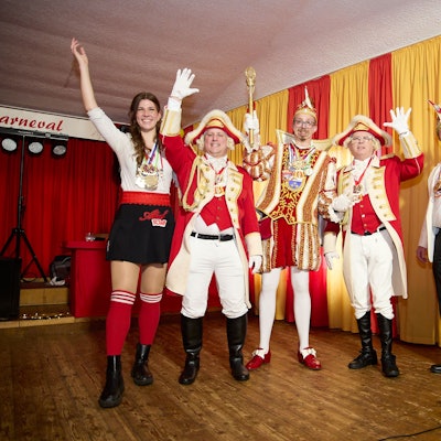 Vier Karnevalisten und eine Karnevalistin stehen auf einer Bühne und werfen jeweils eine Hand in die Luft.