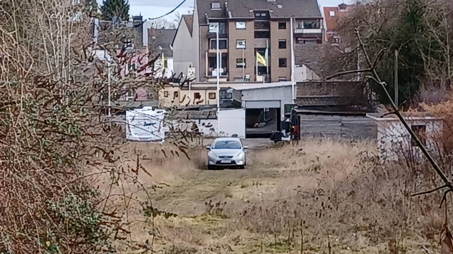 Auf einer Brachfläche steht ein Auto, im Hintergrund sind Häuser zu sehen.