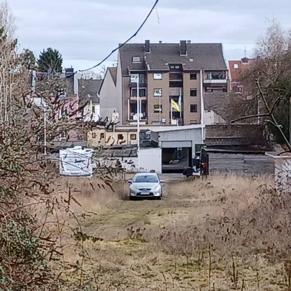 Auf einer Brachfläche steht ein Auto, im Hintergrund sind Häuser zu sehen.