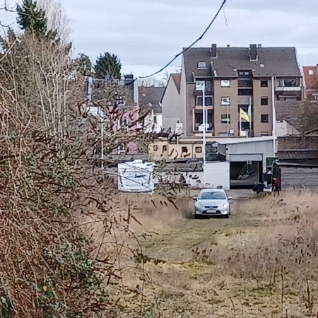 Auf einer Brachfläche steht ein Auto, im Hintergrund sind Häuser zu sehen.