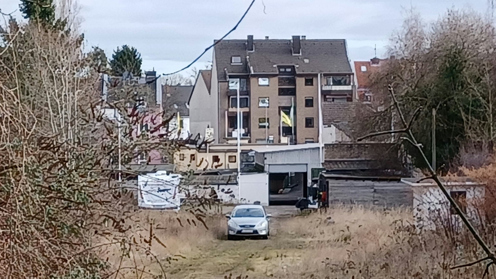 Auf einer Brachfläche steht ein Auto, im Hintergrund sind Häuser zu sehen.