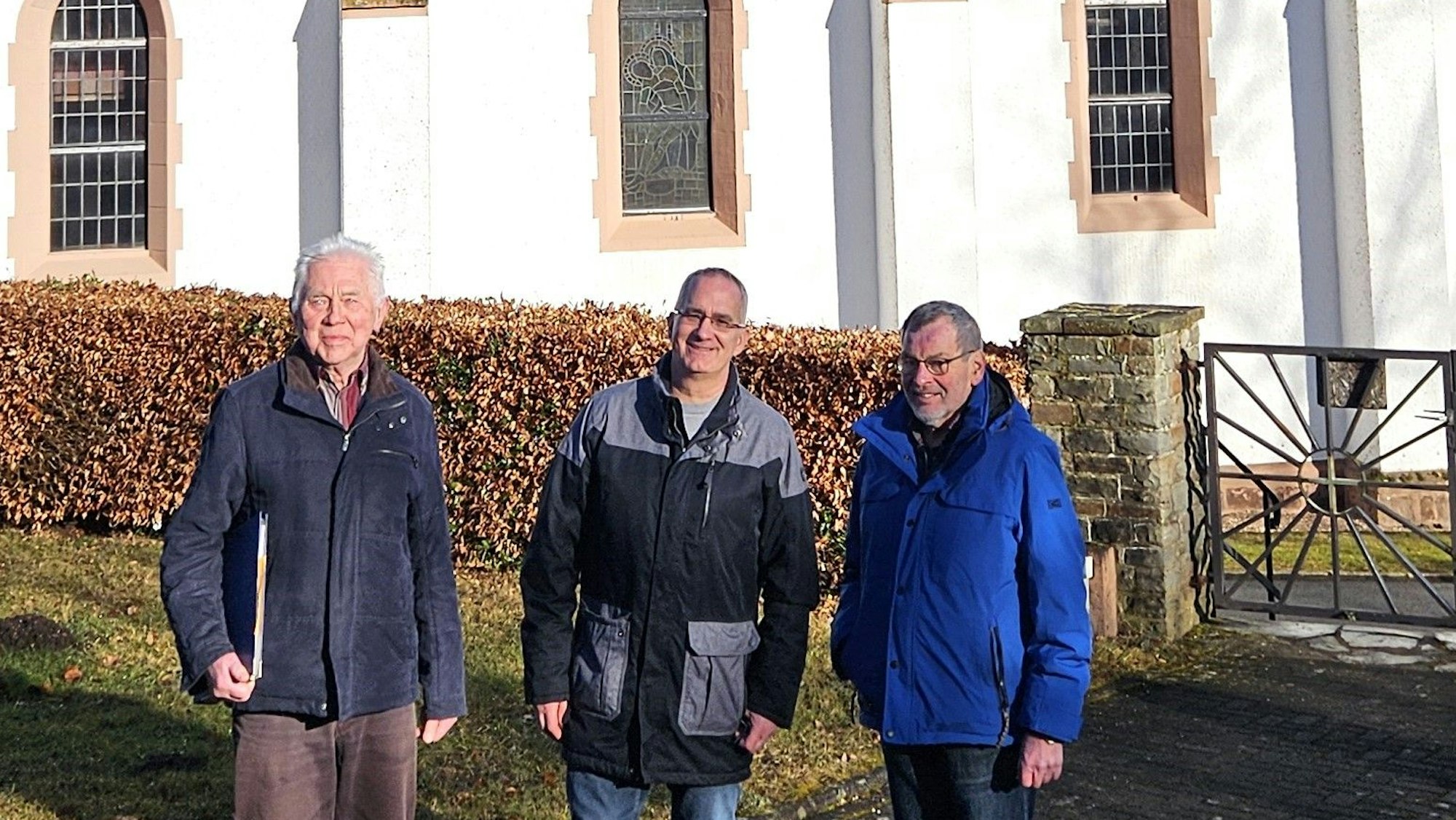 Die drei Männer stehen nebeneinander vor der Kirche.
