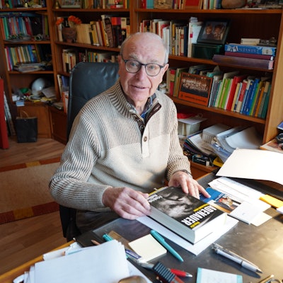 Das Foto zeigt einen älteren Herren an einem Schreibtisch, im Hintergrund stehen Bücher im Regal.