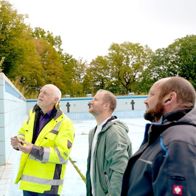 Drei Männer stehen in einem leeren Schwimmbecken und begutachten Baumängel.