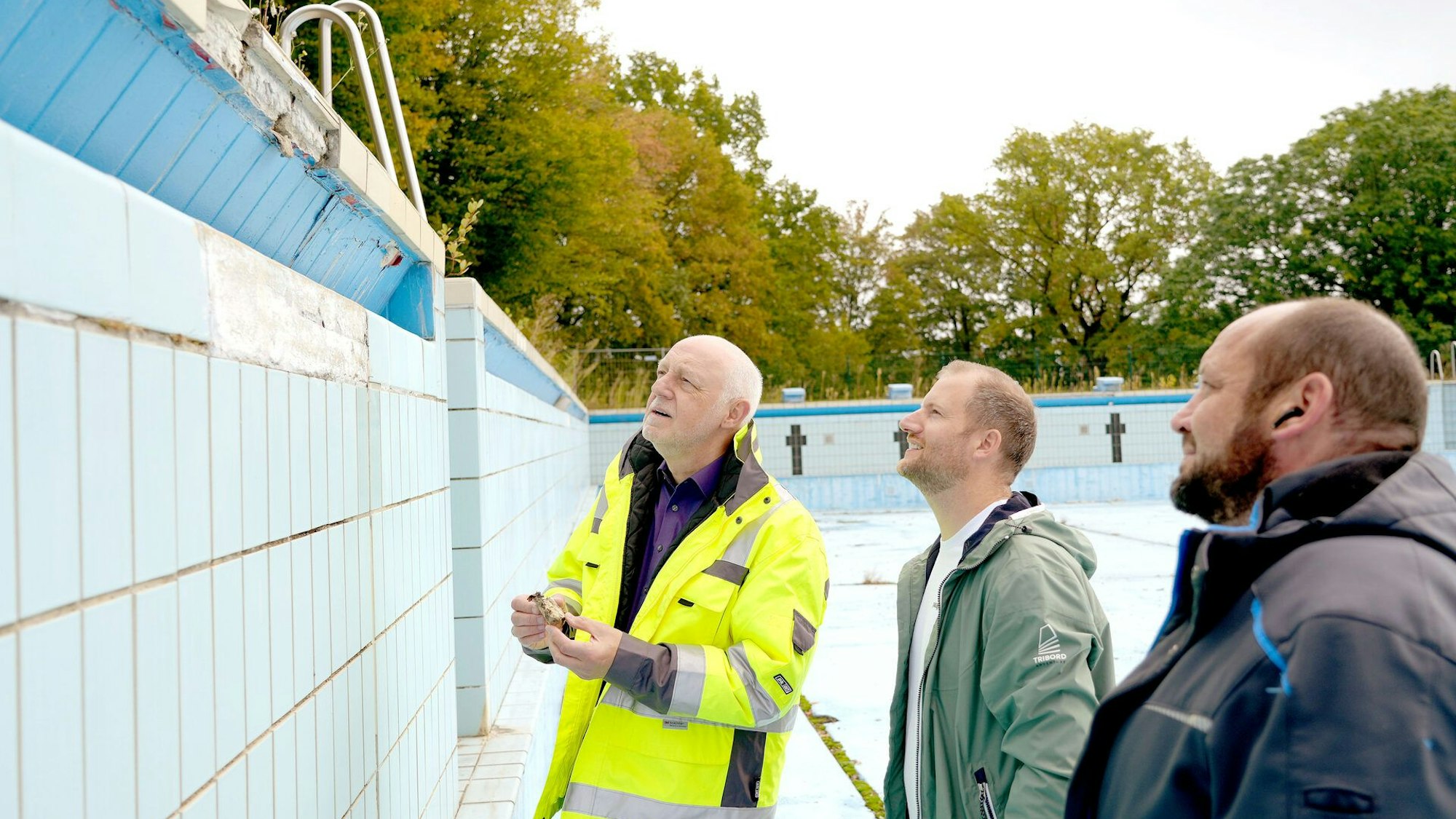 Drei Männer stehen in einem leeren Schwimmbecken und begutachten Baumängel.