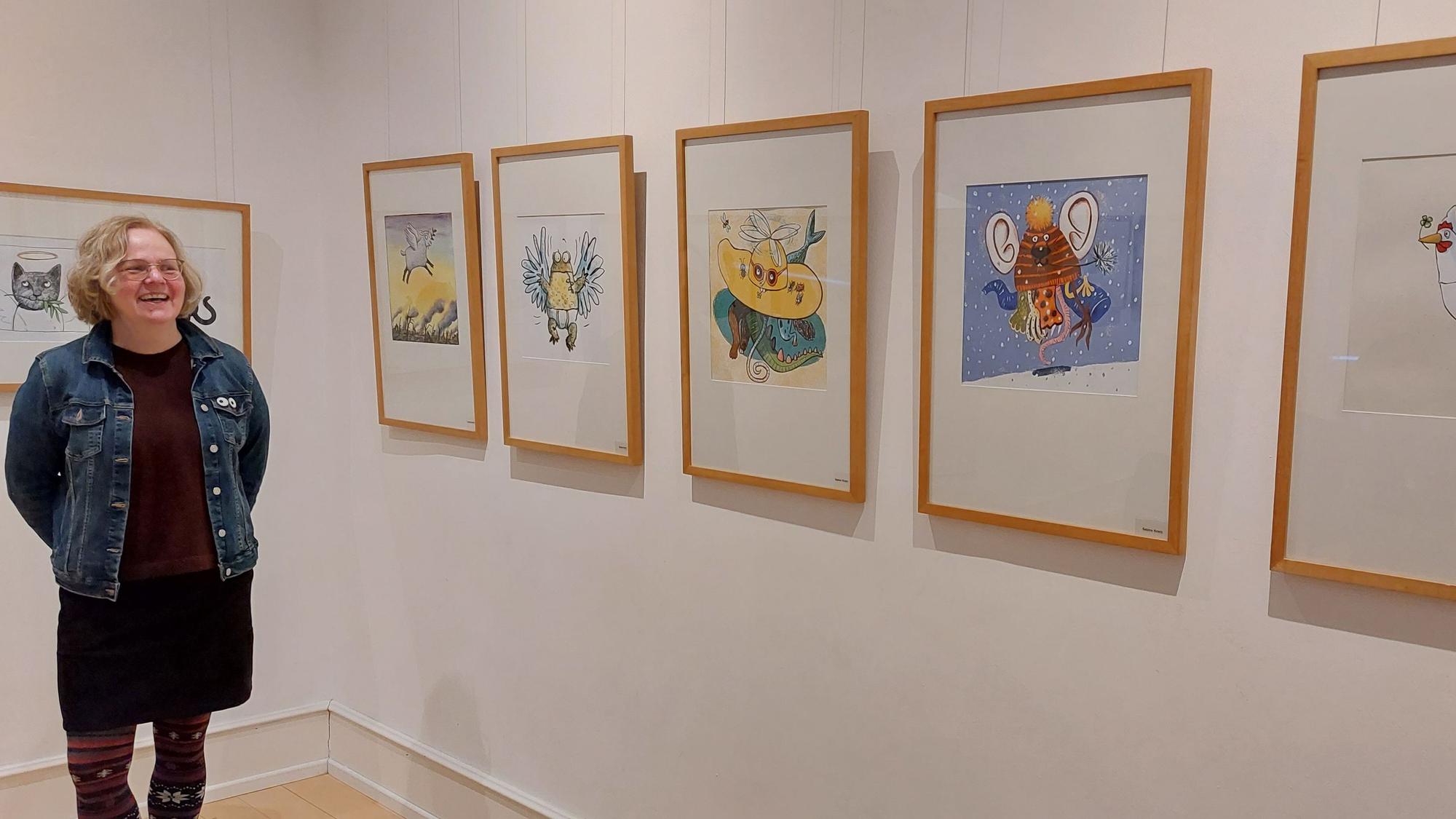 Eine Frau mit blonden Locken steht neben einer Museumswand mit gerahmten Illustrationen.