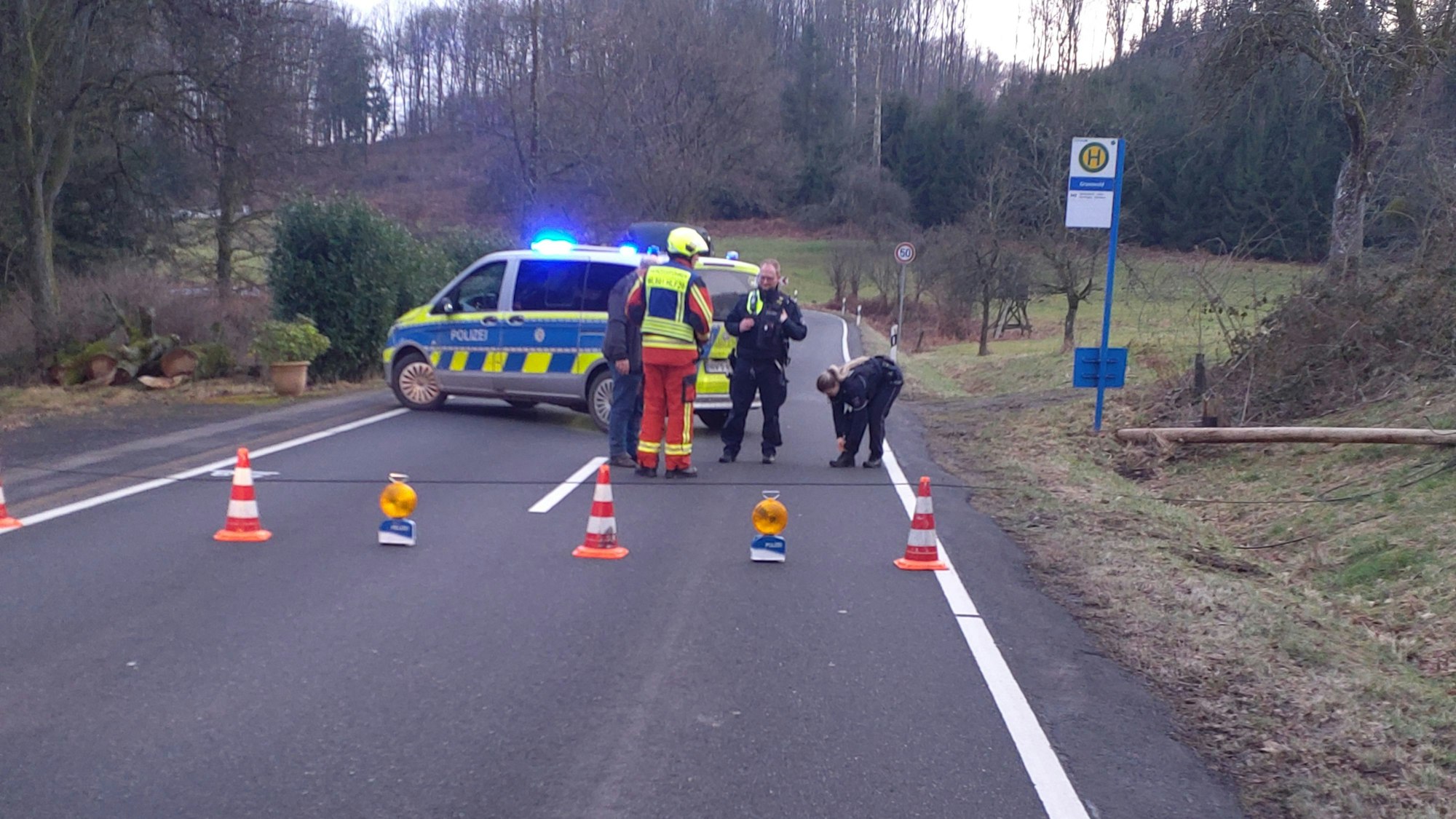 Polizei und Feuerwehr sichern in der Waldbröler Ortschaft die Stelle, an der ein hölzener Kabelmast der Deutschen Telekom umgekippt ist. Der Mast landete im Grünen und nicht auf dem Asphalt der stark befahrenen Bundesstraße 256.