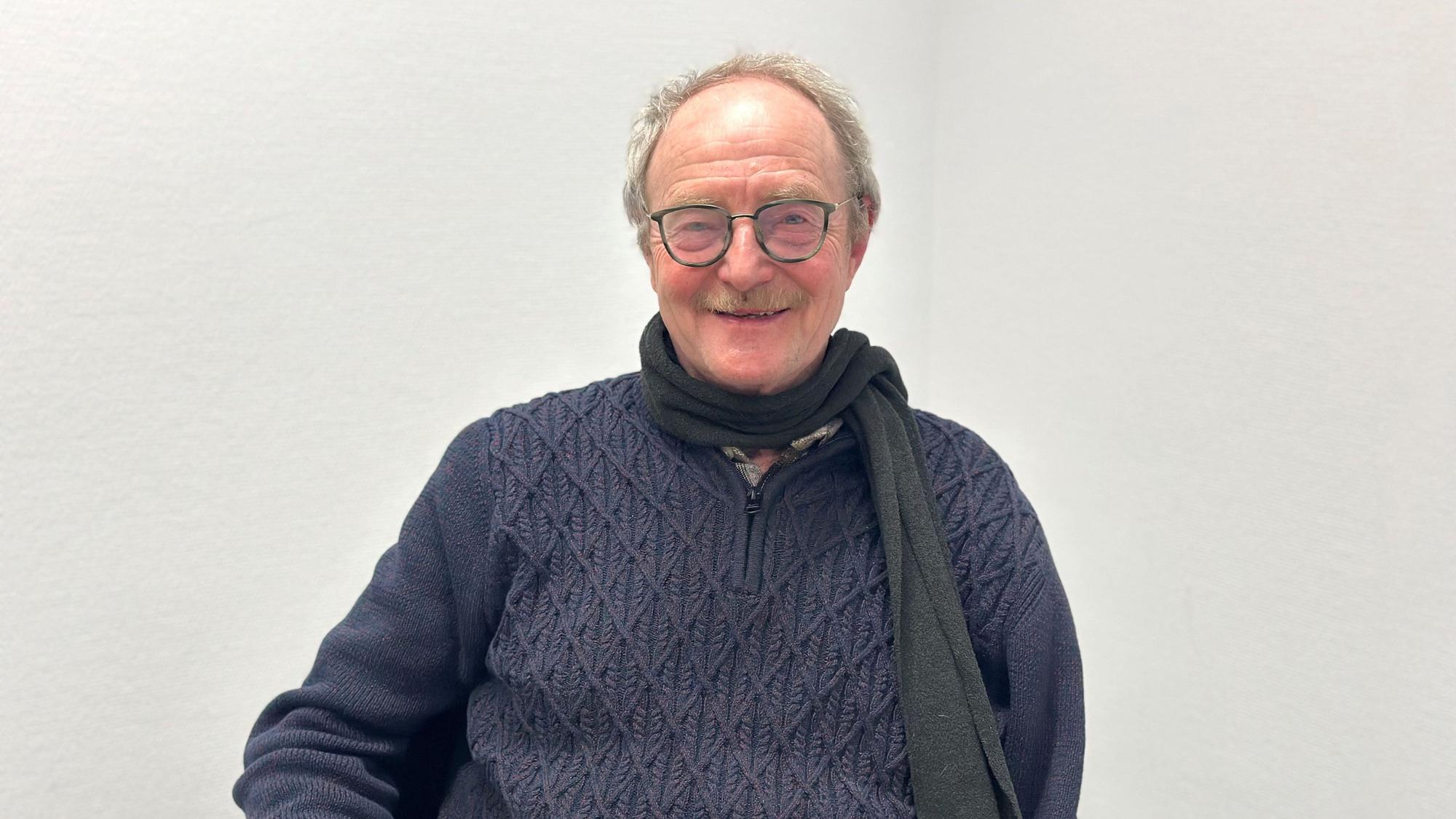 Ein Mann sitzt vor einer weißen Wand und lächelt.