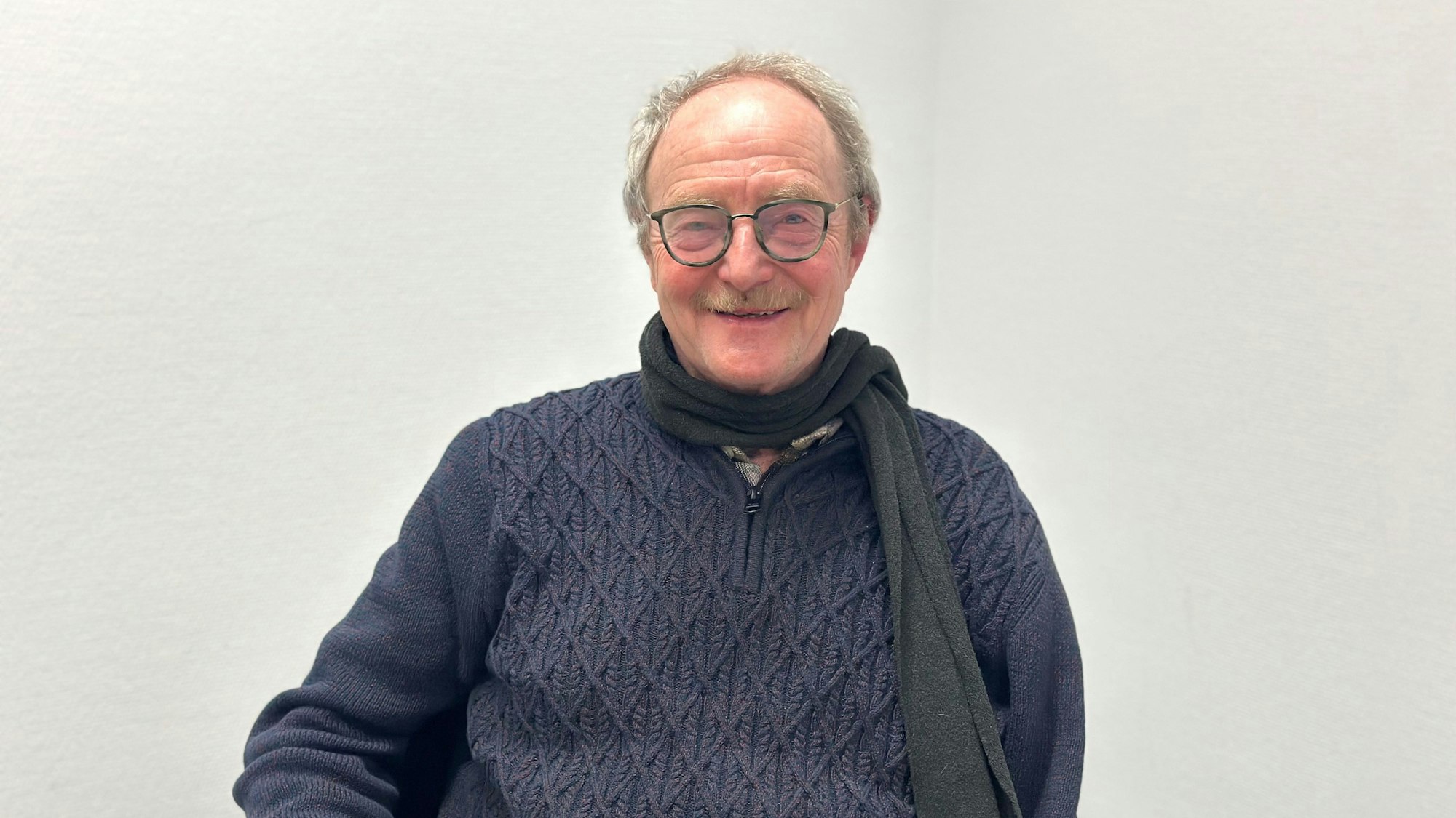 Ein Mann sitzt vor einer weißen Wand und lächelt.