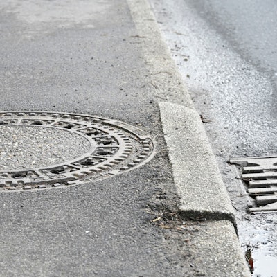 Ein Gehweg mit einem Gullideckel. Ein Stein der Bordsteinkante hat sich gesenkt.