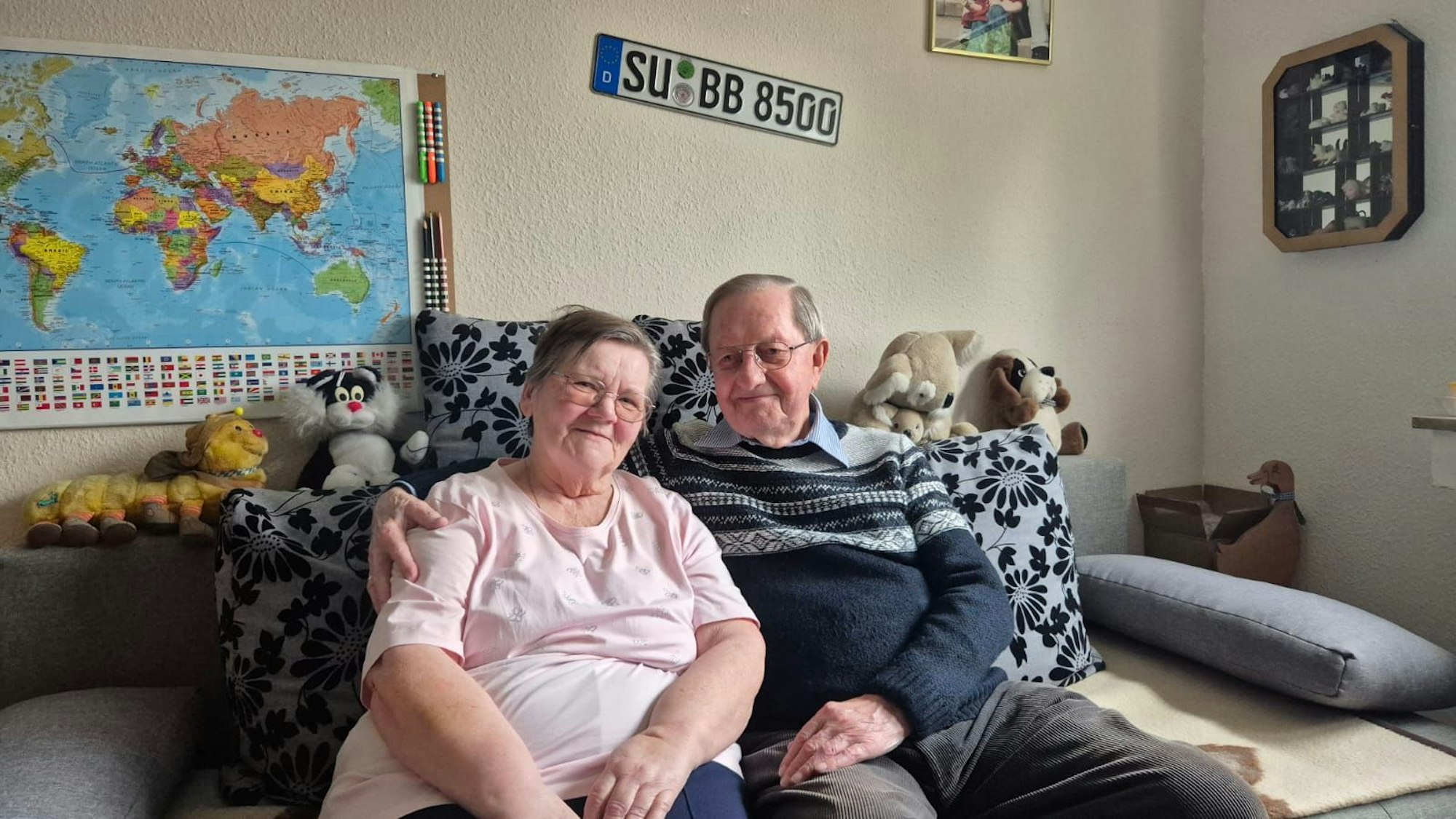 Ein älteres Ehepaar sitzt auf einem Sofa, auf dessen Lehne Kuscheltiere sitzen.
