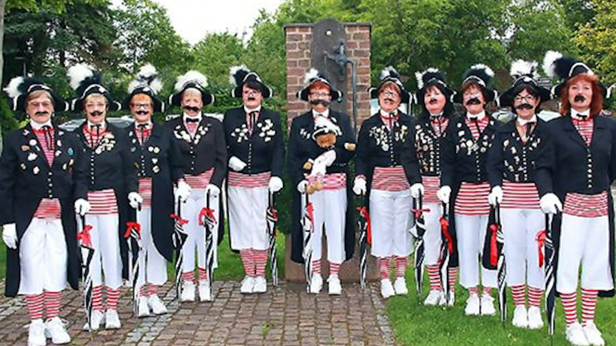 Eine Gruppe in rotweißen Ringelshirts, schwarzen Jacken und weißen Hosen steht nebeneinander. Alle haben angeklebte Schnäuzer.