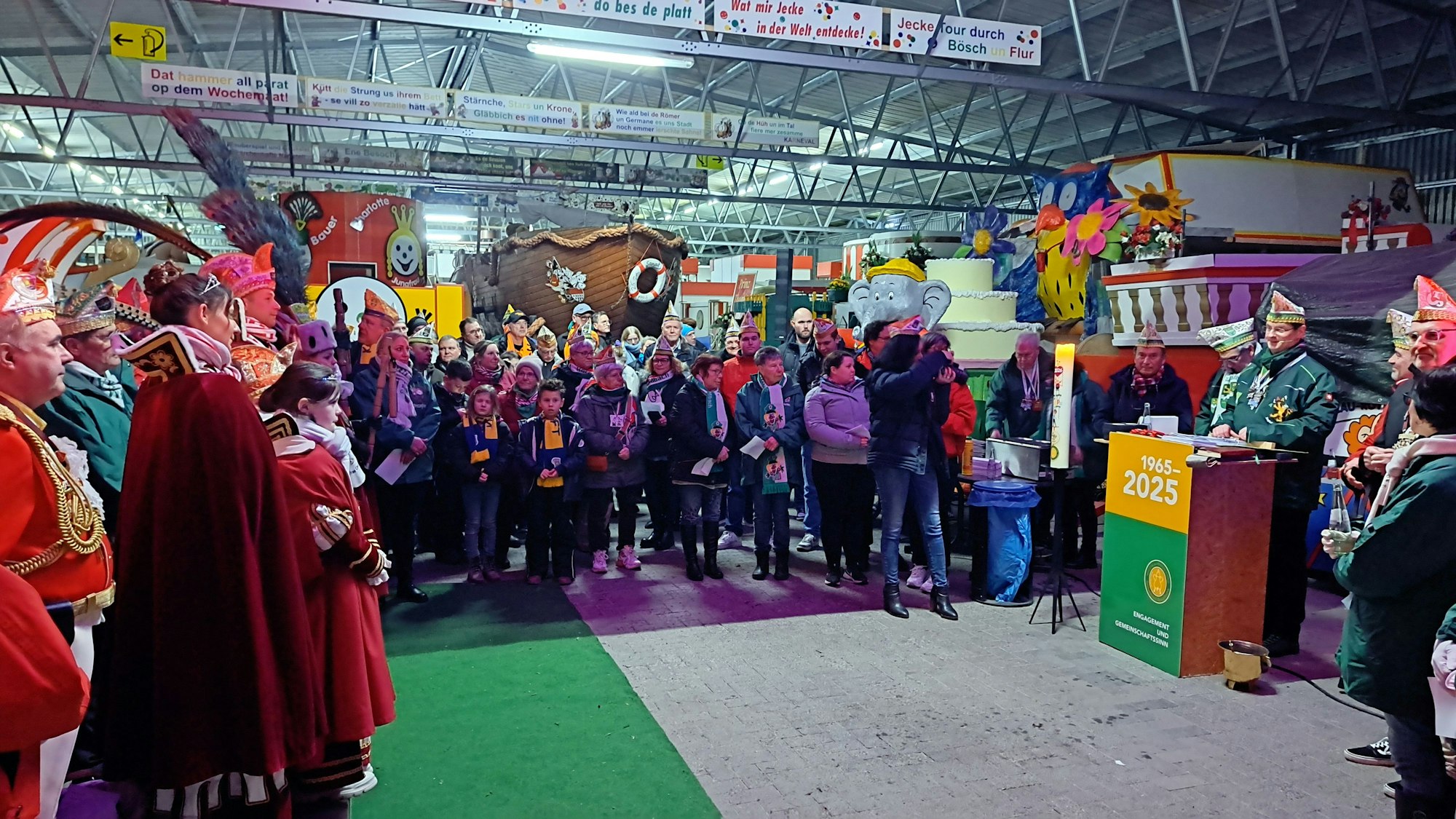 Viele Karnevalisten stehen in einer halle, in der verschiedene Karnevalswagen aufgebaut sind, darunter ein Piratenschiff.