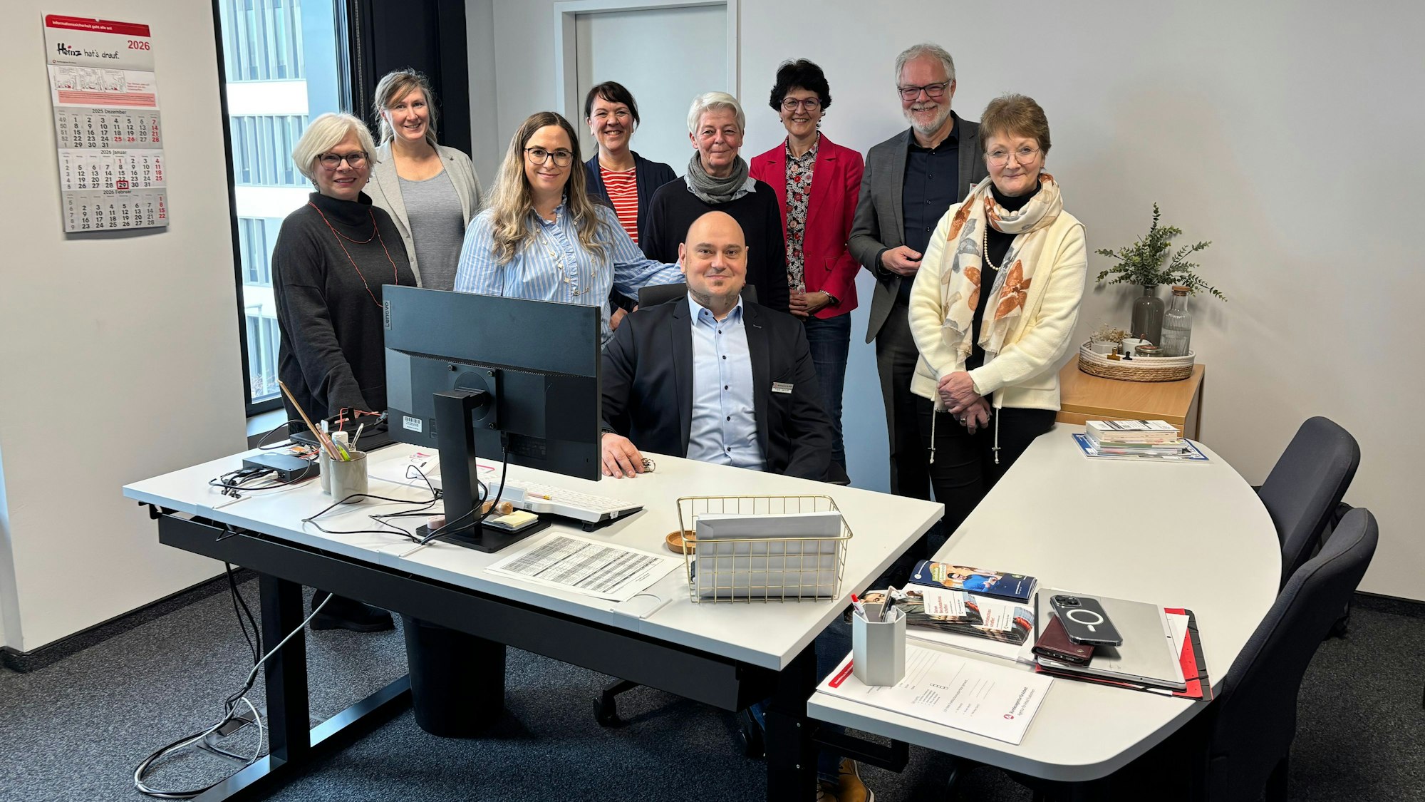 Eine Gruppe Menschen steht hinter Schreibtischen in einem Büroraum. Ein Mann sitzt vor ihnen und hat seine Hand auf einem der Schreibtische abgestützt.