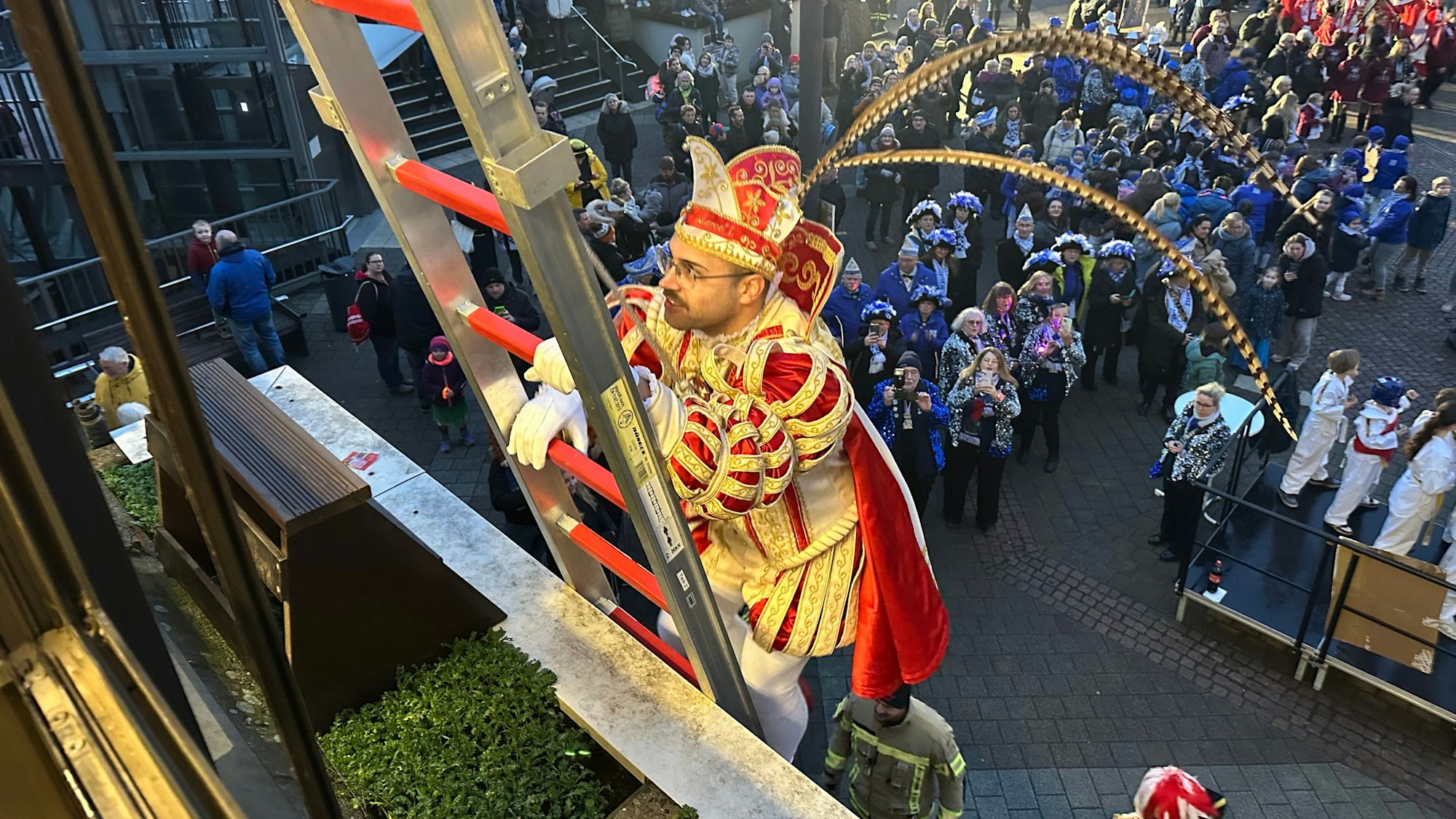 Ein Karnevalsprinz steigt eine Leiter hinauf, während unten kostümierte Garden zuschauen.