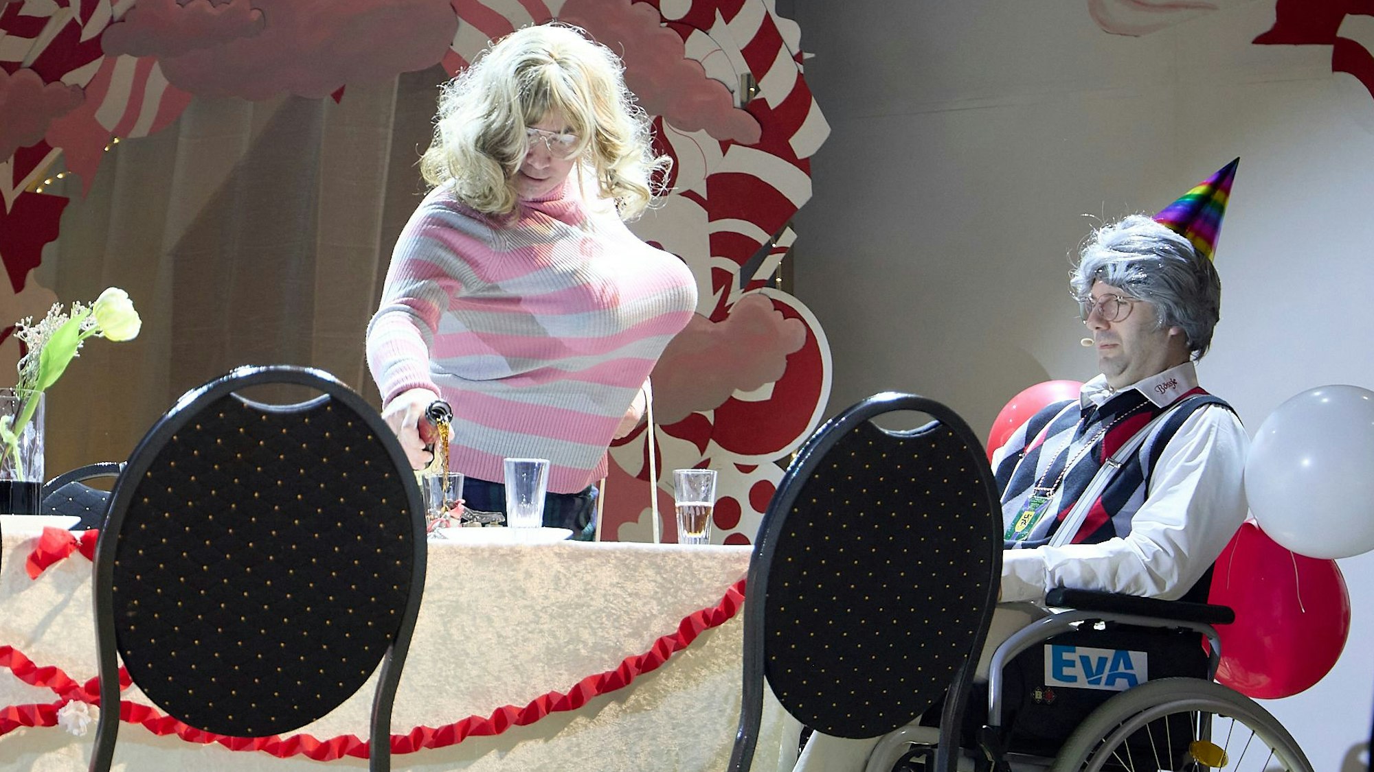 Ein Mann, der überzeichnet als Frau verkleidet ist, gießt Schnaps in ein Glas ein. Am Tisch sitzt ein weiterer Mann in einem Rollstuhl. Er trägt eine Altherrenperrücke.