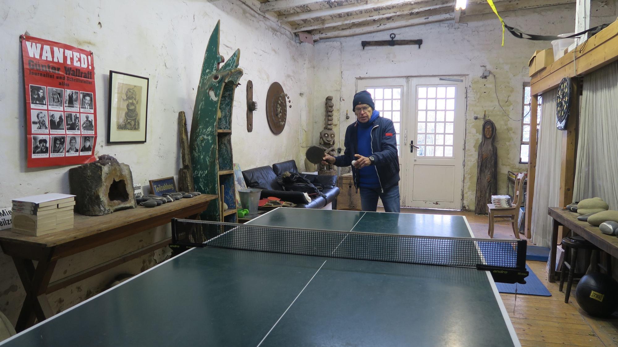 Ein Mann mit blauer Mütze, blauer Jacke und Jeans steht an einer Tischtennisplatte, bereit zum Aufschlag.