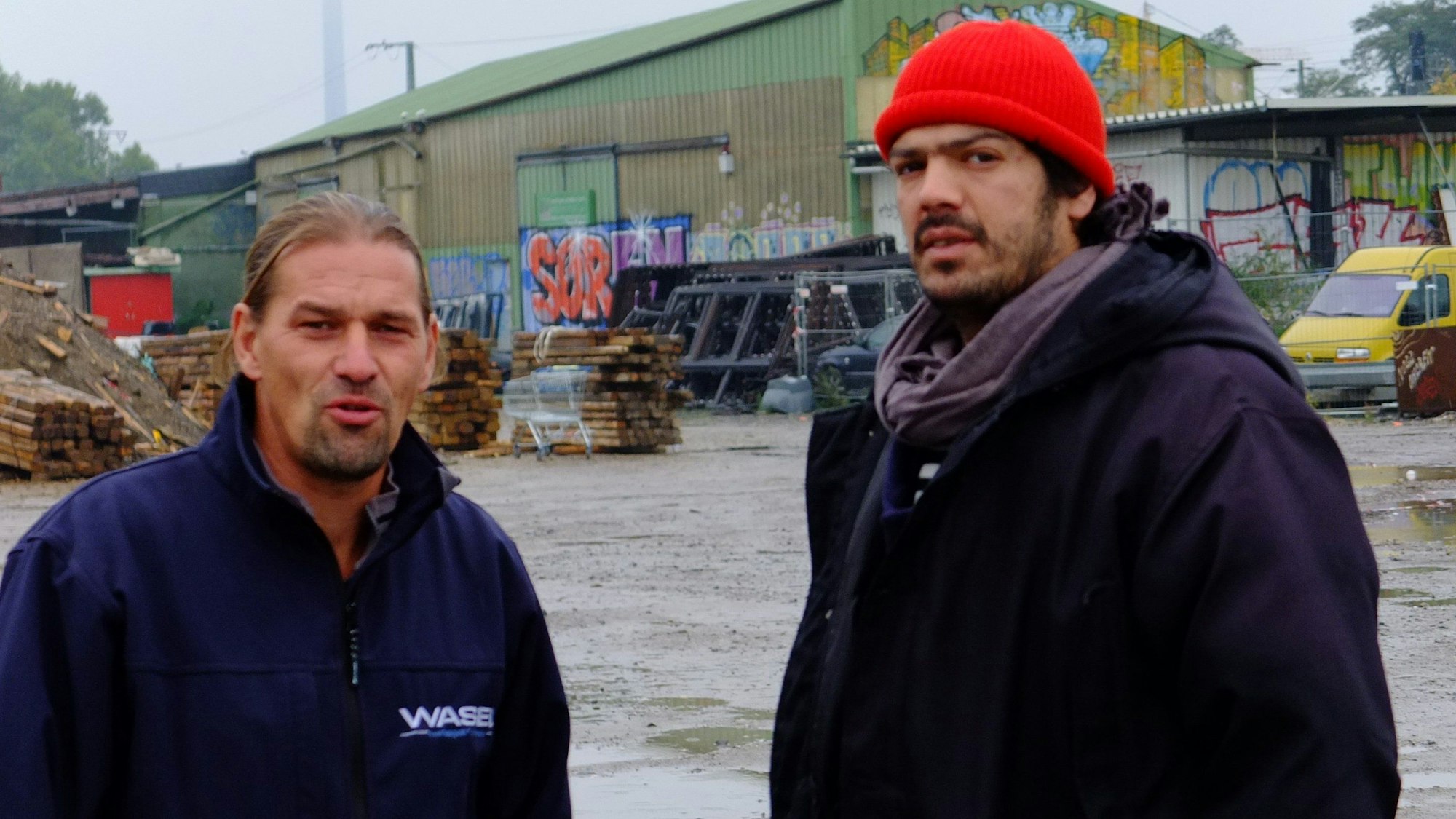 Zwei Männer stehen vor einer großen grünen Lagerhalle auf einem schlammigen Boden.