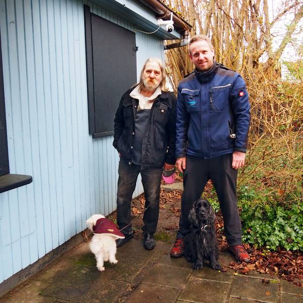 Zwei Männer stehen mit ihren beiden Hunden neben der Laube einer Kleingartenanlage.