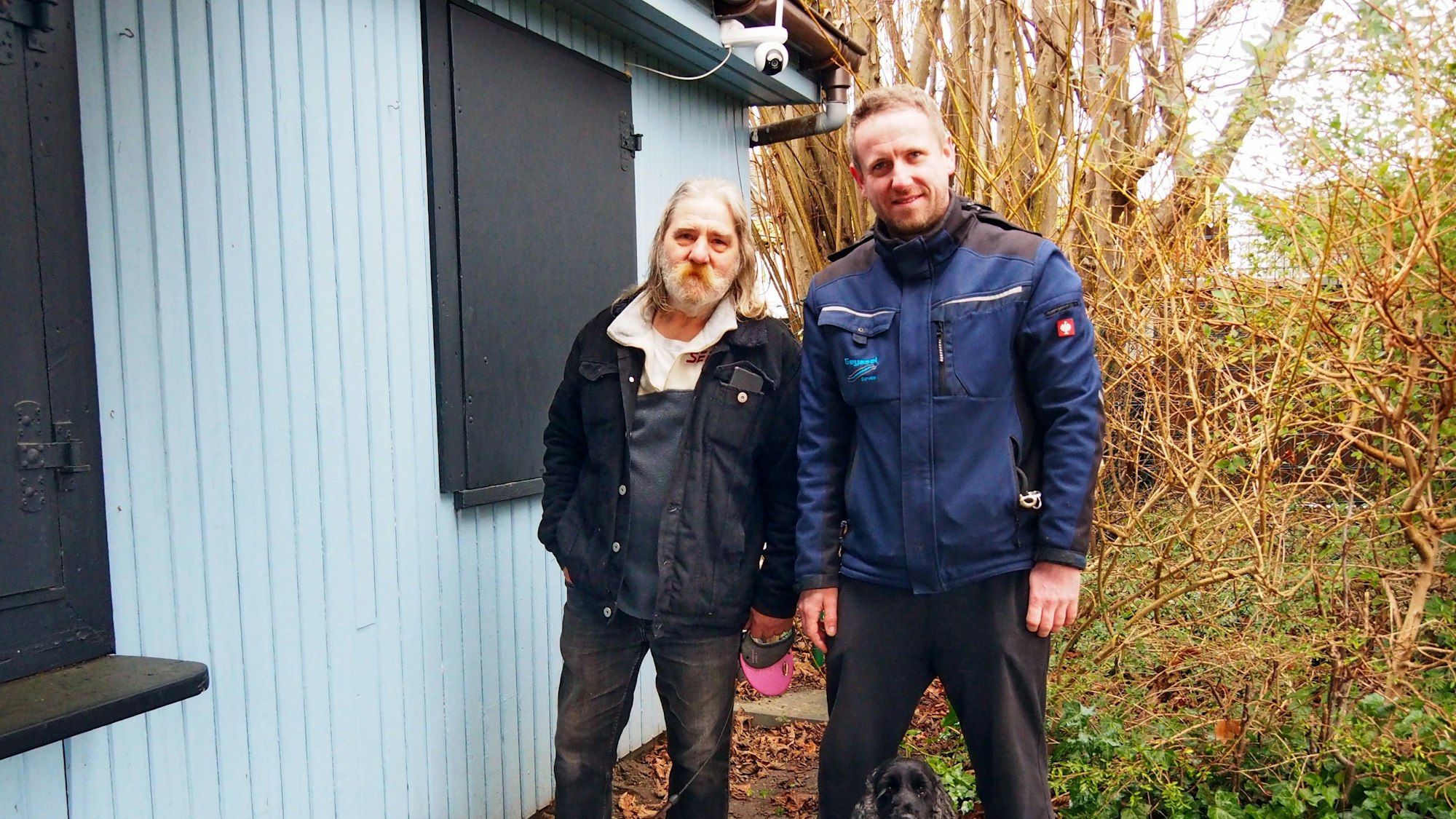 Zwei Männer stehen mit ihren beiden Hunden neben der Laube einer Kleingartenanlage.