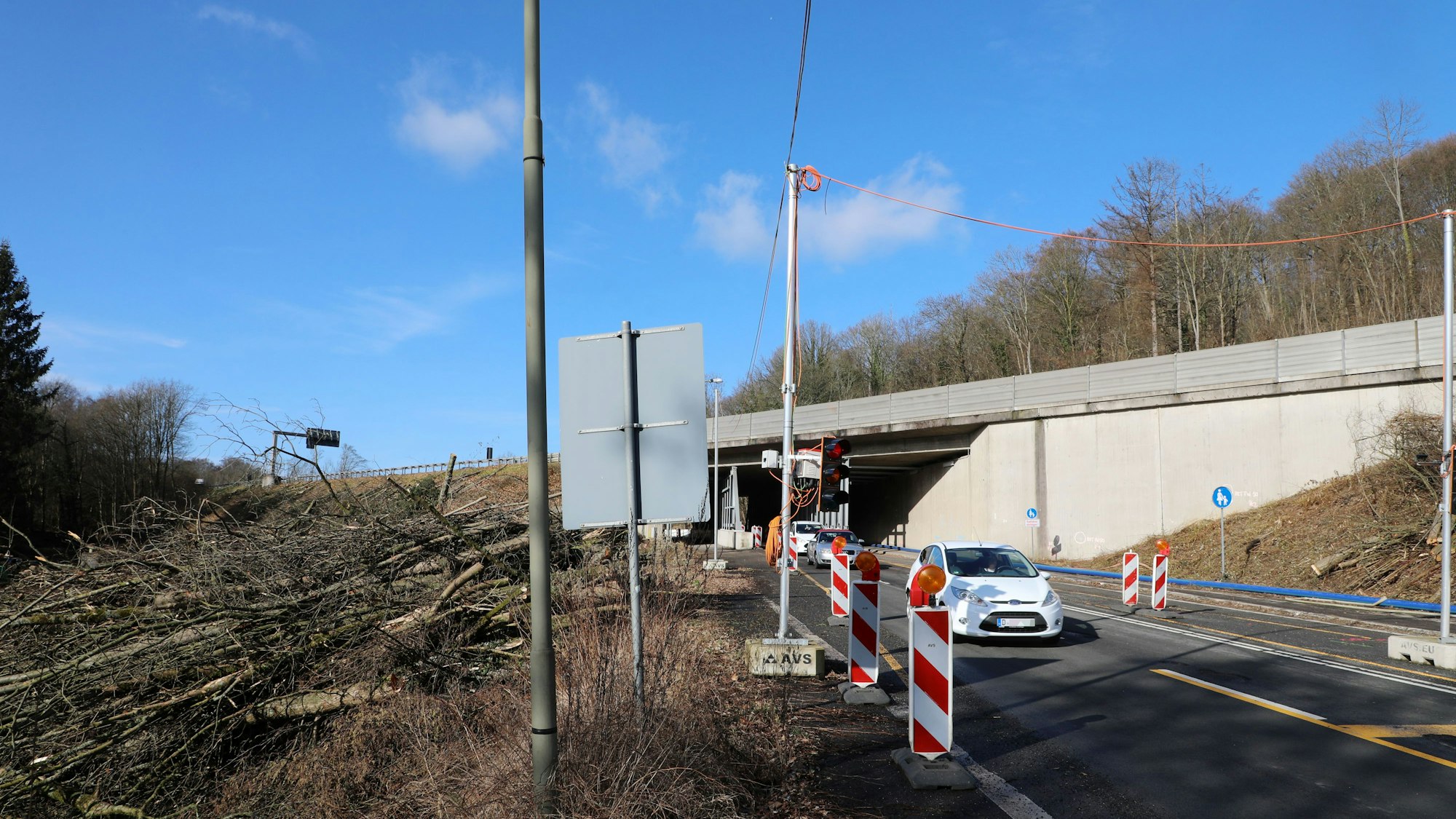links Baustämme auf dem Boden liegend, rechts ein weißes Auto, das gerade eine Unterführung durchfahren hat.