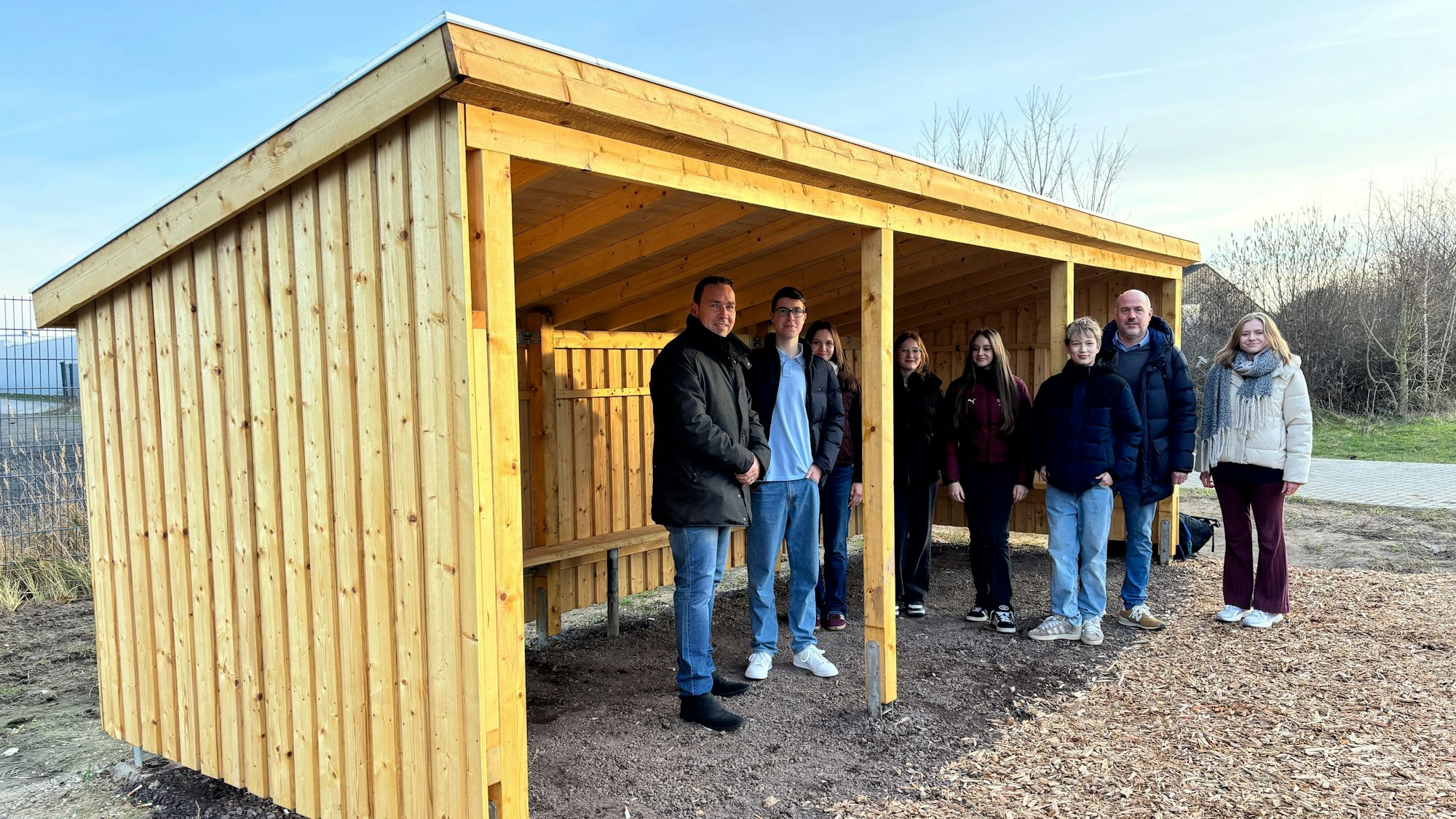 Eine Gruppe Erwachsener und Jugendlicher steht unter und neben einem hölzernen Unterstand.