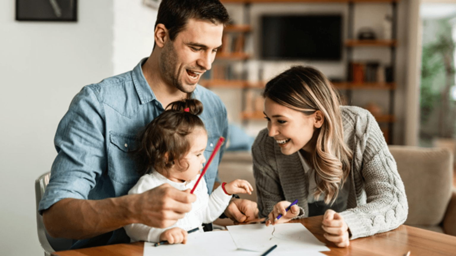 Auf dem Bild sitzt eine kleine Familie fröhlich zusammen am Tisch und malt mit bunten Stiften. Die Erwachsene links und rechts unterstützen das Kind liebevoll beim Malen und lachen gemeinsam.