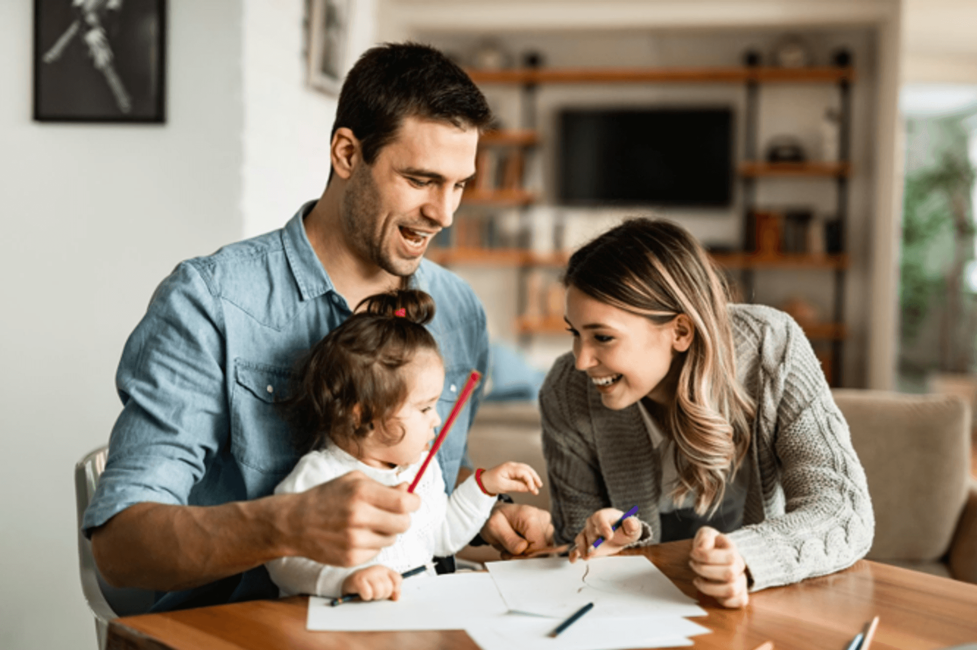 Auf dem Bild sitzt eine kleine Familie fröhlich zusammen am Tisch und malt mit bunten Stiften. Die Erwachsene links und rechts unterstützen das Kind liebevoll beim Malen und lachen gemeinsam.