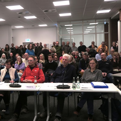 An einem Tisch sitzen sechs Menschen, dahinter ist Publikum auf Stühlen und stehend zu sehen.