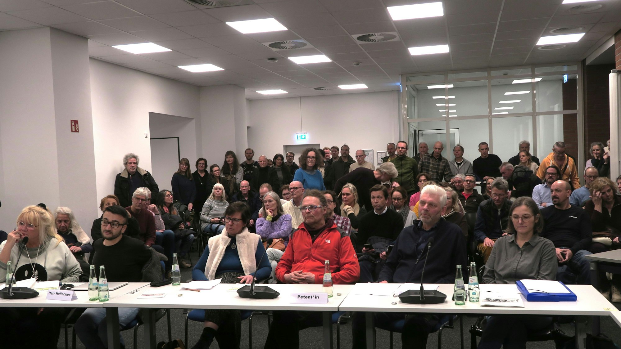 An einem Tisch sitzen sechs Menschen, dahinter ist Publikum auf Stühlen und stehend zu sehen.
