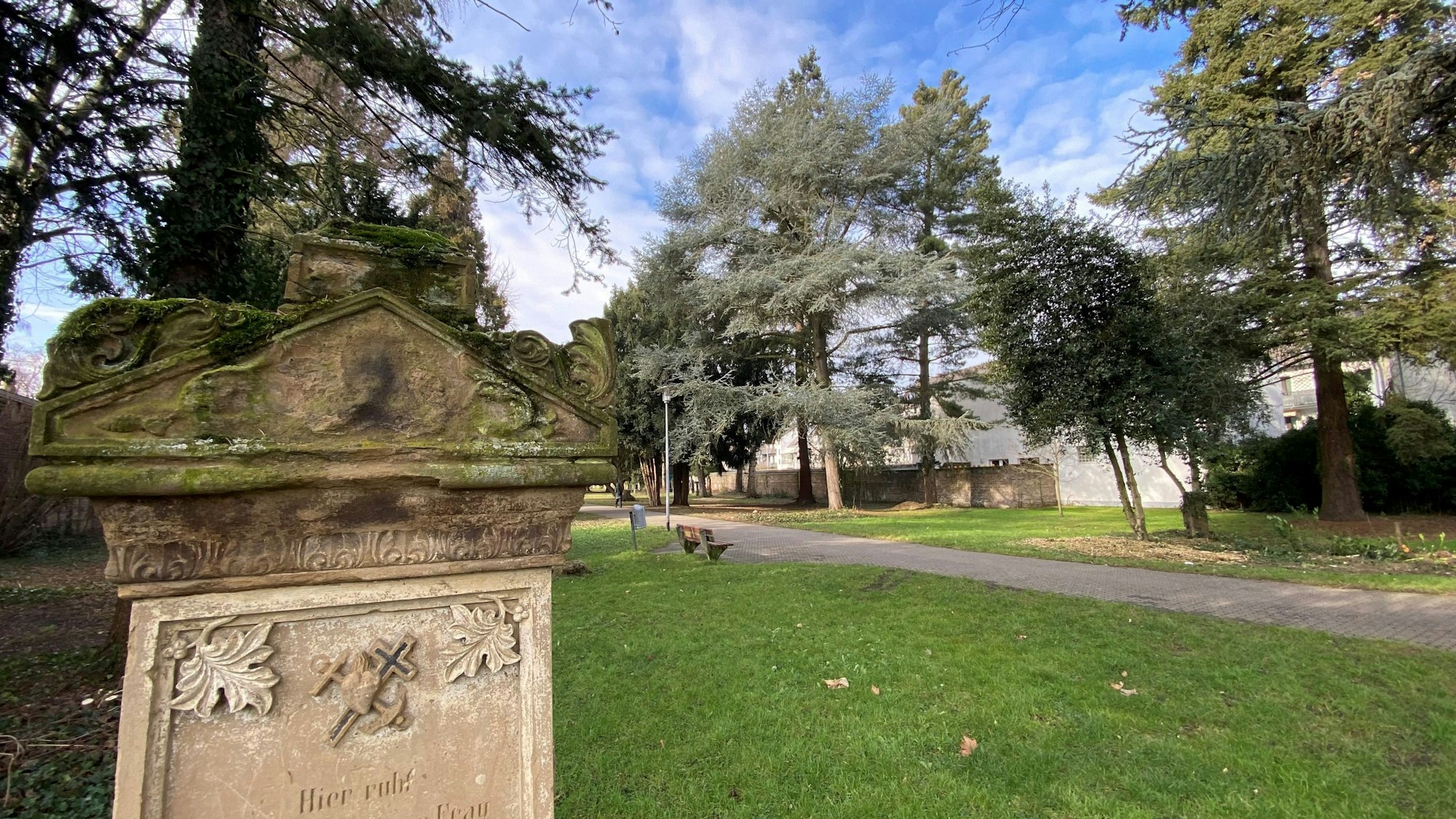 Zu sehen sind ein Grabstein und im Hintergrund ein Park.