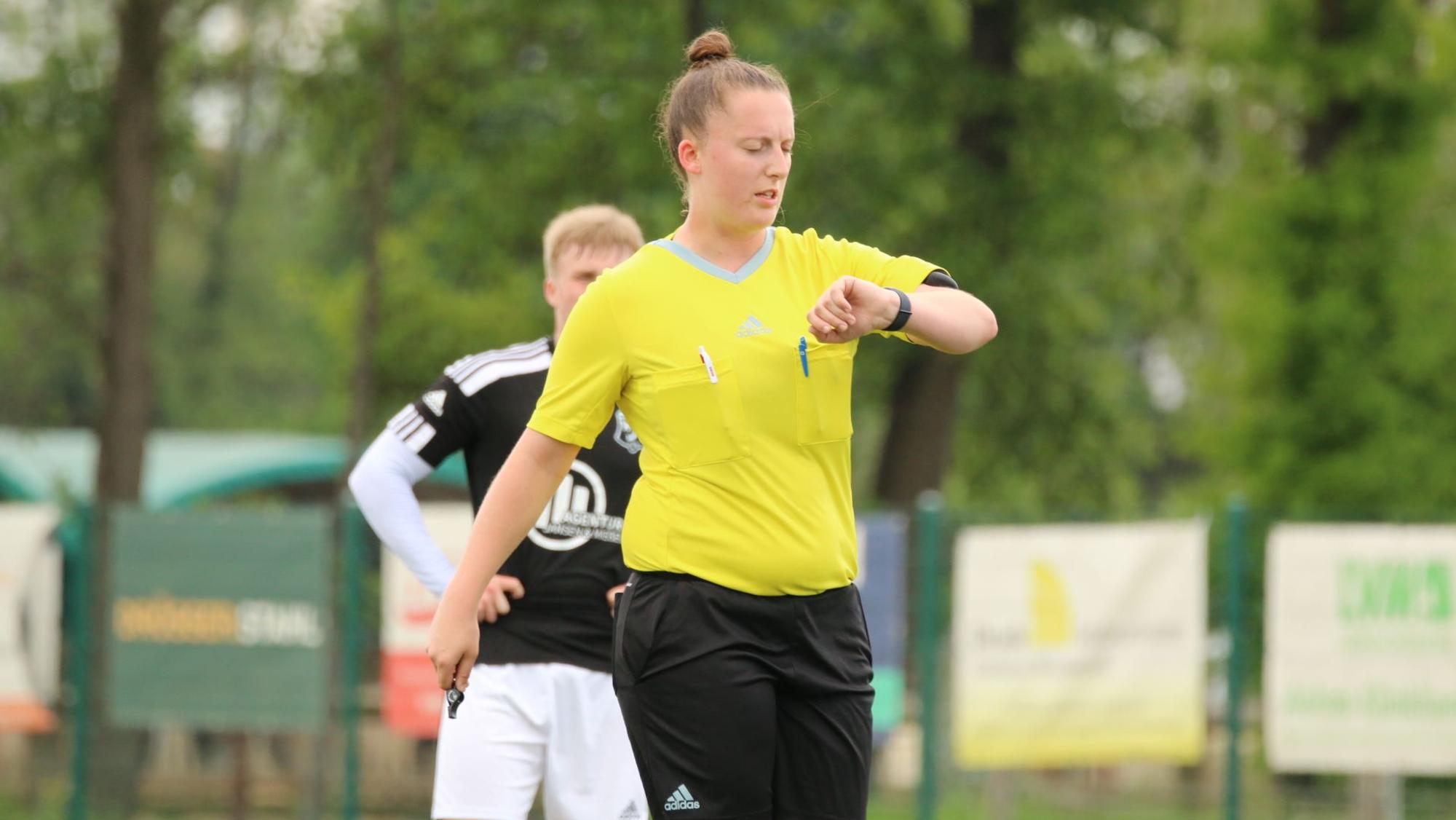 Eine Frau steht in einem gelben T-Shirt und schwarzer Sporthose auf einem Fußballplatz und schaut auf ihre Uhr.