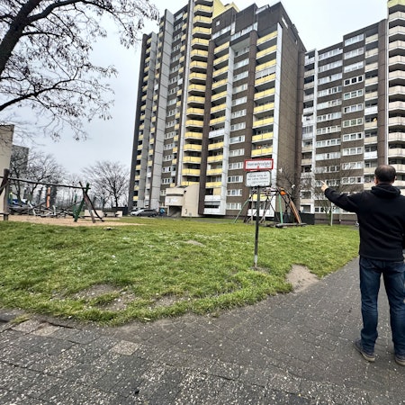 Ein Mann steht mit dem Rücken zur Kamera, er steht vor einer Grünanlage vor drei gewaltigen Hochhäusern.