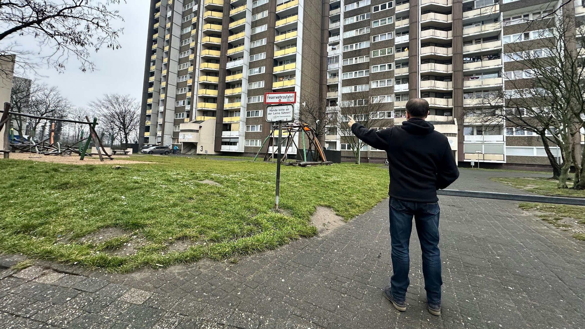 Ein Mann steht mit dem Rücken zur Kamera, er steht vor einer Grünanlage vor drei gewaltigen Hochhäusern.