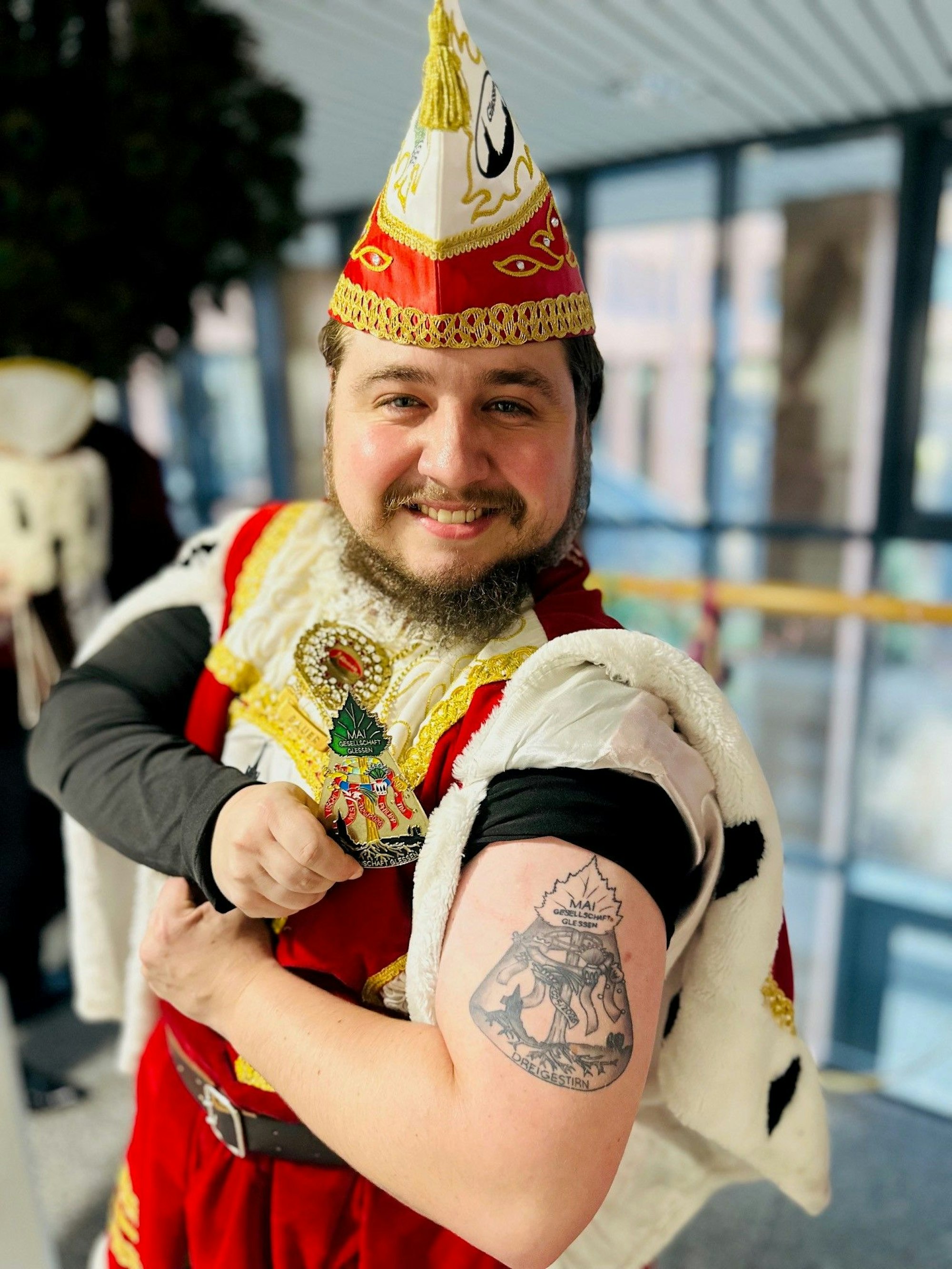 Das Foto zeigt einen Mann im Ornat, der eine Tätowierung auf dem Oberarm präsentiert.