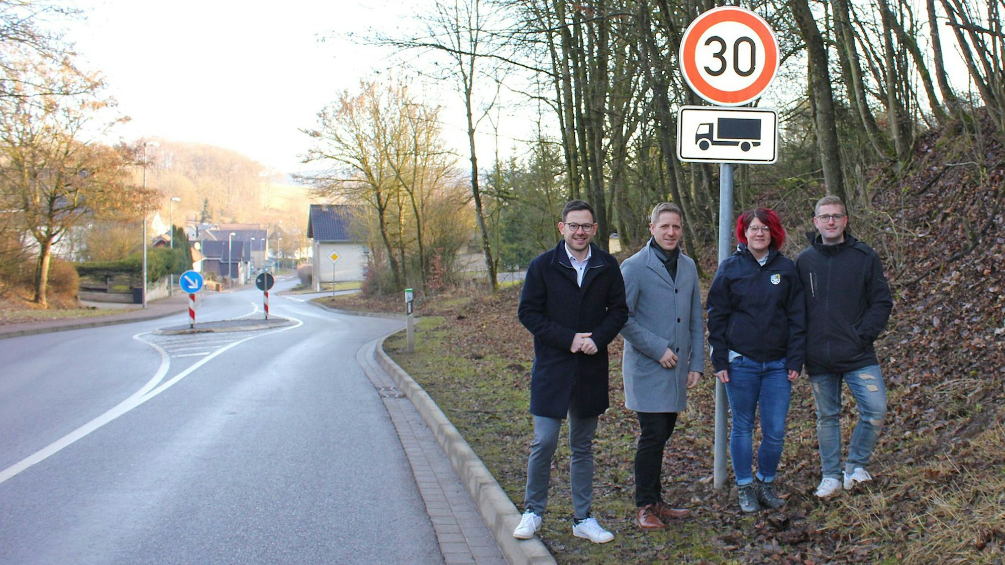 Drfei Männer und eine Frau stehen neben einer Landstraße an zwei Schildern, die auf Tempo 30 für Lkw hinweisen.