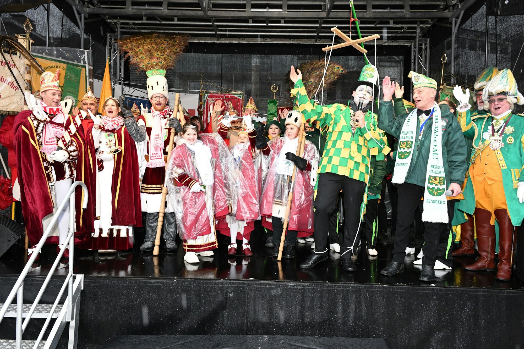 Das Erwachsenen- und das Kinder-Dreigestirn stehen auf einer Bühne neben einem Kostümierten, an dessen Arme und Beine Fäden befestigt sind, die zu einem Marionettenkreuz gehören.