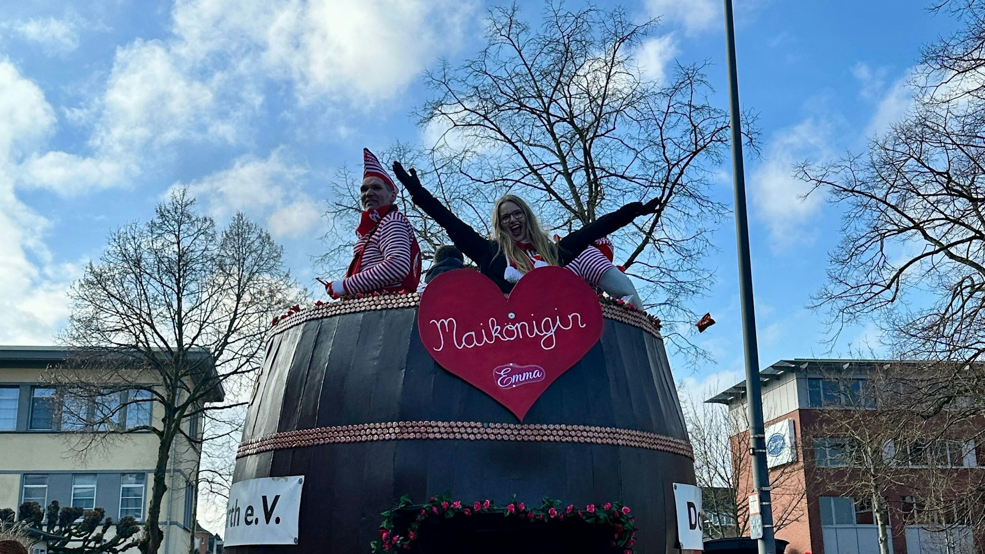 Aus einem Karnevalswagen, der wie ein riesiges Fass gebaut ist, schaut oben die Maikönigin heraus.