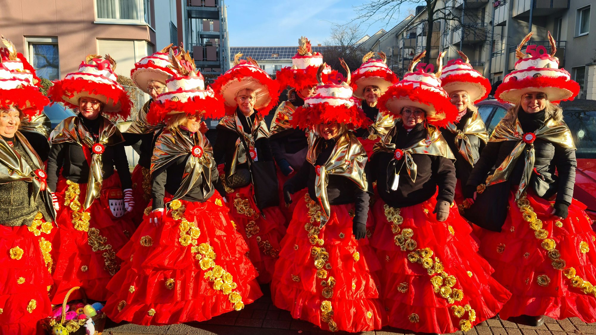 Eine Gruppe Frauen mit riesigen Tortengleichen Hüten und roten Röcken mit goldenen Rosen darauf stehen nebeneinander.