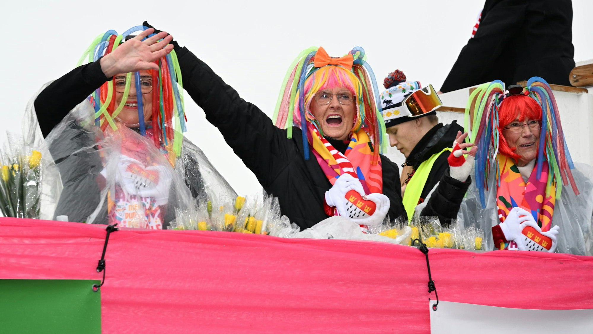 Drei Frauen mit Perücken in den Farben des Regenbogens stehen auf einem geschmückten Karnevalswagen und winken.