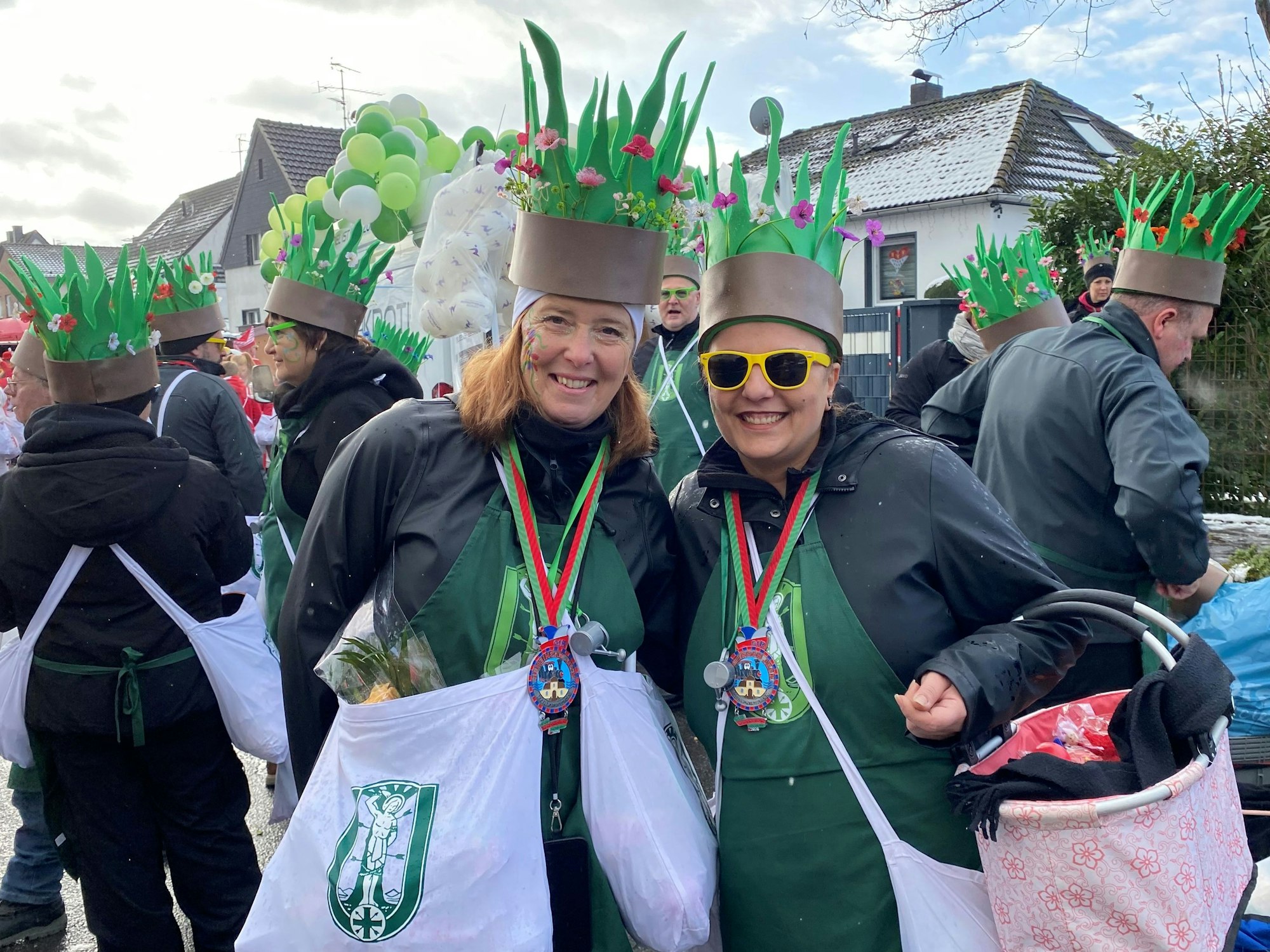 Zwei Frauen mit grünen Schürzen. Auf dem Kopf - aus Pappe - grünes Gras und bunte Blumen.